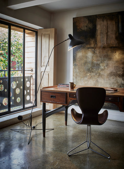A modern home office scene featuring a dark brown molded leather swivel chair, a wooden writing desk, a black floor lamp, and a large textured abstract wall painting.