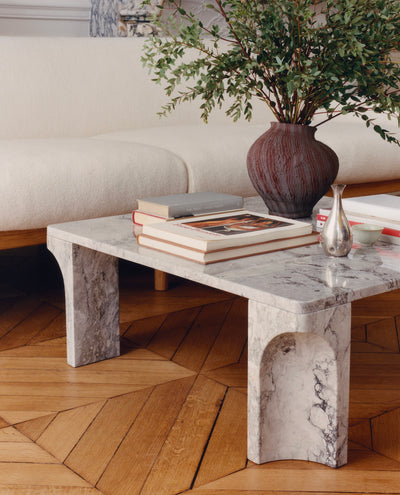 A modern marble coffee table with chunky block legs and a curved lower panel, styled in a living room setting with books, a vase, and a sofa in the background.