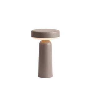 A minimalist table lamp with a cylindrical base and a circular top, shown in a matte taupe-gray color with a warm light glowing from the middle, set against a solid white background.