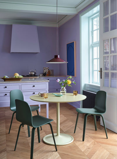A dining area in a modern kitchen with a round cream table and four dark green chairs, set against lavender purple walls and light wood herringbone flooring.
