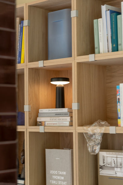 A modern black table lamp with a glowing illuminated shade sits on a wooden bookshelf filled with books and decor.