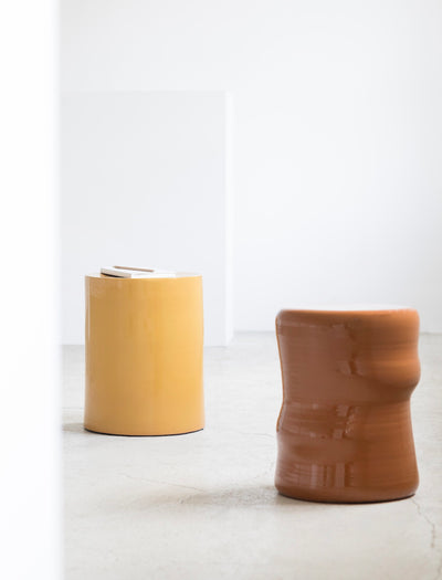 Two modern ceramic stools in a minimalist studio setting. One is a smooth, glossy mustard yellow cylinder, and the other is a textured, rustic terracotta brown stool with a wavy, organic silhouette.